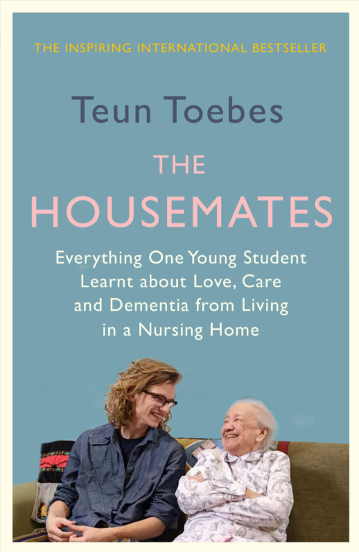 Book cover of 'The Housemates' by Teun Toebes with two people sitting on a couch against a blue background.