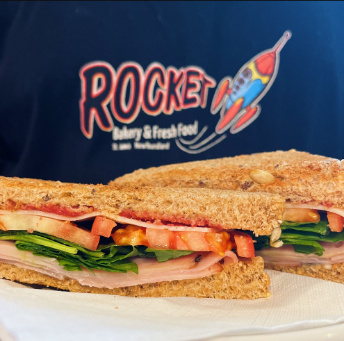 Half-cut sandwich with visible ingredients on a plate in front of Rocket Bakery & Fresh Food logo.